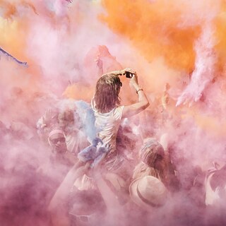 Menschenmenge bei einem Festival, umgeben von farbigem Pulver in Rosa‑, Orange‑ und Gelbtönen; eine Person wird über der Menge hochgehoben und hält die Arme nach oben.