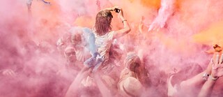 Menschenmenge bei einem Festival, umgeben von farbigem Pulver in Rosa‑, Orange‑ und Gelbtönen; eine Person wird über der Menge hochgehoben und hält die Arme nach oben.