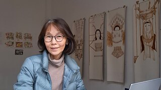 An Asian woman working at a desk with a laptop, surrounded by drawings.