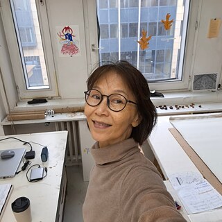 Eine Frau mit Brille lächelt, während sie ein Selfie vor einem Schreibtisch mit verschiedenen Gegenständen darauf macht.