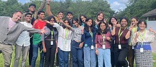 Group of PASCH students standing outdoors, wearing name badges and posing together in front of a green background.
