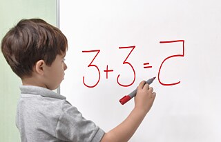Ein Junge mit einer dyslexischen Beeinträchtigung schreibt das Ergebnis einer Rechenaufgabe auf ein Whiteboard. Seitenansicht.