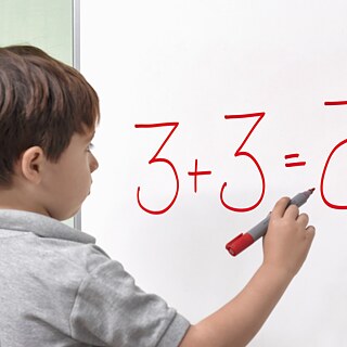 Ein Junge mit einer dyslexischen Beeinträchtigung schreibt das Ergebnis einer Rechenaufgabe auf ein Whiteboard. Seitenansicht.