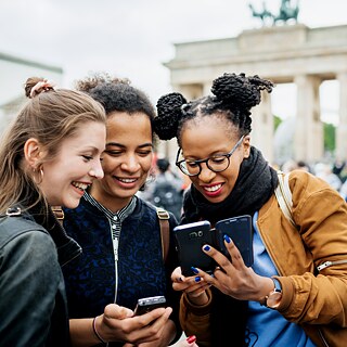 Drei junge Frauen stehen vor dem Brandenburger Tor in Berlin und betrachten lachend die Fotos auf einem Smartphone.