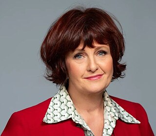 Porträt von Annette Dittert, die ein rotes Jackett über einer hellen, gemusterten Bluse trägt, vor einem schlichten grauen Hintergrund.