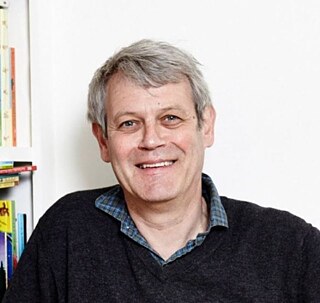 Portrait of Axel Scheffler wearing a dark jumper over a checkered shirt, standing against a white background. A bookshelf with colourful books is visible on the left.
