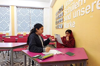 Zwei junge Frauen sitzen an einem Tisch und unterhalten sich, auf dem Tisch liegen Arbeitsblätter, eine Brille und Schreibwerkzeug.