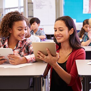 Eine Lehrerin hockt zwischen zwei Schülerbänken. In der Hand hält sie ein Tablet. Auf dem erklärt sie gerade einer Schülerin etwas. Im Hintergrund sieht man weitere Schülerinnen und Schüler, die an ihren Bänken etwas auf dem Tablet machen.