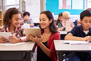 Eine Lehrerin hockt zwischen zwei Schülerbänken. In der Hand hält sie ein Tablet. Auf dem erklärt sie gerade einer Schülerin etwas. Im Hintergrund sieht man weitere Schülerinnen und Schüler, die an ihren Bänken etwas auf dem Tablet machen.