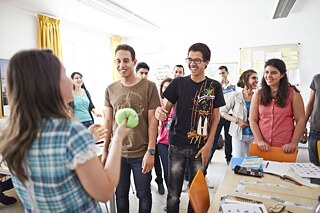 A group of people is standing in the classroom. A person is standing in front of them. The person is interacting with the students and holding teaching materials.