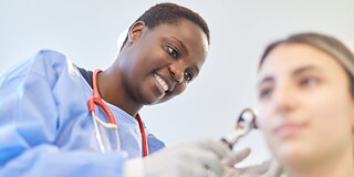 A young medical student wearing a blue lab coat and a stethoscope is examining a seated patient's right ear. The background is very blurry.