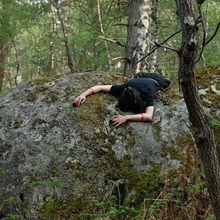 Eine Person liegt bäuchlings über einem großen, moosbedeckten Felsblock in einem dichten Wald. Die Arme hängen seitlich über den Stein herab, umgeben von Bäumen, Farnen und Waldboden. Das Foto zeigt eine ruhige Waldszene mit natürlichen Farben und weichem Licht. 