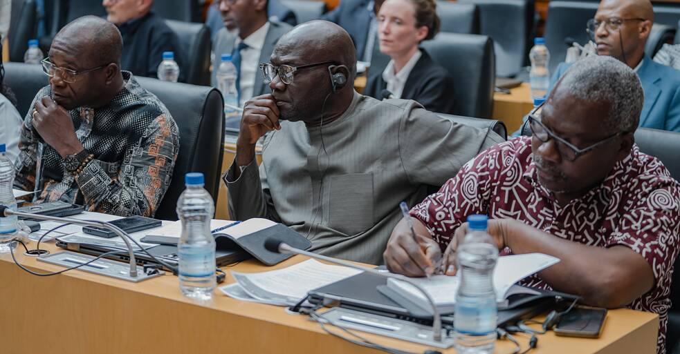 (AU Commission, attentive delegates) Three African museum directors seated at the conference table in the AU Commission chamber, listening attentively to the proceedings. The central figure wears traditional grey attire and a translation earpiece; the man to his right takes notes in a notebook. Delegates are visible in the rows behind them.