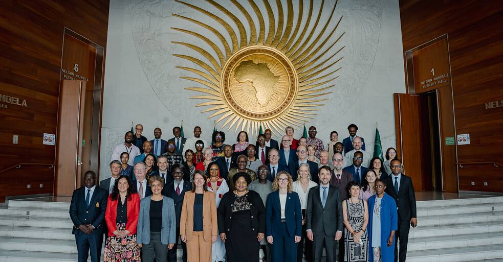 (AU Commission, group photograph) A large group of approximately 50 people posed for a formal photograph in front of the African Union Commission's iconic golden sunburst emblem. The Nelson Mandela Plenary Hall doors are visible on either side. Participants from across Africa and Europe stand together on the steps.