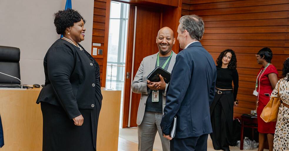 (AU Commission, networking) Three people in conversation in a formal wood-panelled room at the African Union Commission. A woman in a black suit stands facing two men — one in a grey blazer with a green conference lanyard, smiling warmly; the other in a navy suit with his back to the camera. An AU flag is visible in the background, alongside other guests.
