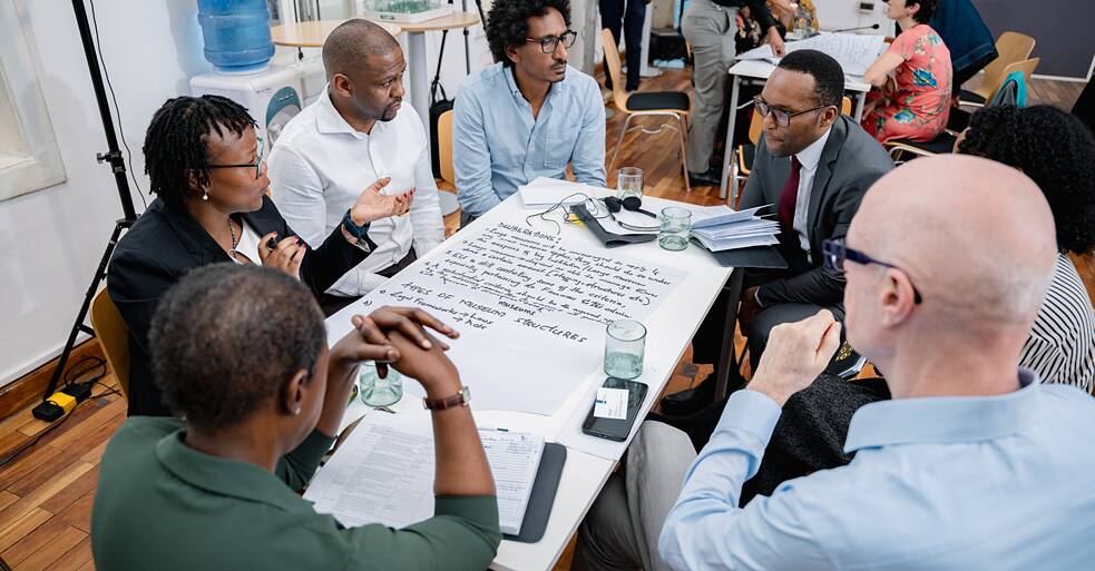 (Workshop, working group) An overhead view of six museum professionals seated around a table covered in large sheets of paper with handwritten notes. The words "DELIBERATIONS" and "TYPES OF MUSEUM STRUCTURES" are visible on the paper. The room is busy with other groups working in the background.