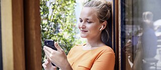 A person with headphones is looking at their smartphone at the window, with green plants in the background.