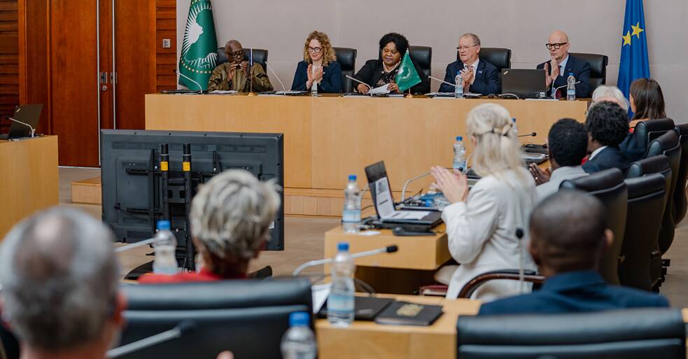 Fünf Podiumsteilnehmende sitzen am erhöhten Podium im Plenarsaal der Afrikanischen Union und applaudieren. Die Flaggen der Afrikanischen Union und der Europäischen Union stehen auf beiden Seiten. Das Publikum, im Vordergrund mit dem Rücken zur Kamera, applaudiert ebenfalls.