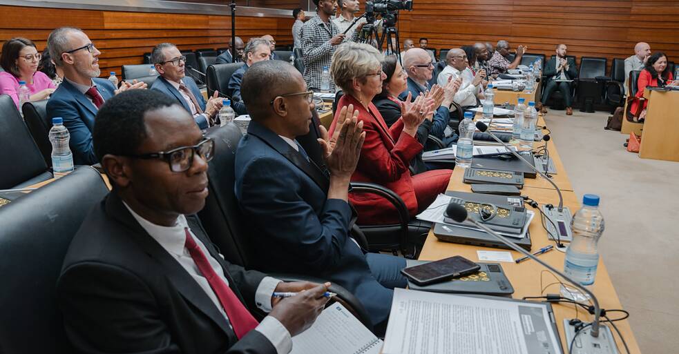 Weitwinkelaufnahme von Delegierten, die an einem langen Konferenztisch im Plenarsaal der Afrikanischen Union sitzen und applaudieren. Mikrofone, Wasserflaschen und Programmdokumente sind auf dem Tisch zu sehen. Kameraleute stehen im hinteren Teil des Raumes.