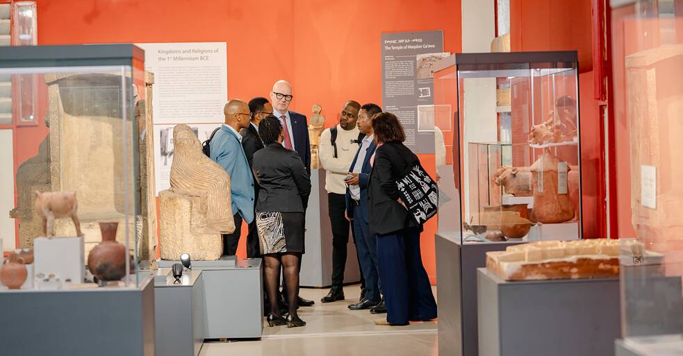 Eine kleine Gruppe von sechs Personen im Gespräch zwischen Vitrinen in einem orangefarbenen Ausstellungsraum des Nationalmuseums von Äthiopien. Antike Steinskulpturen und Keramiken sind in den Vitrinen zu sehen. Eine teilnehmende Person trägt eine bedruckte Tragetasche; die Gruppe ist in einen informellen Austausch vertieft.
