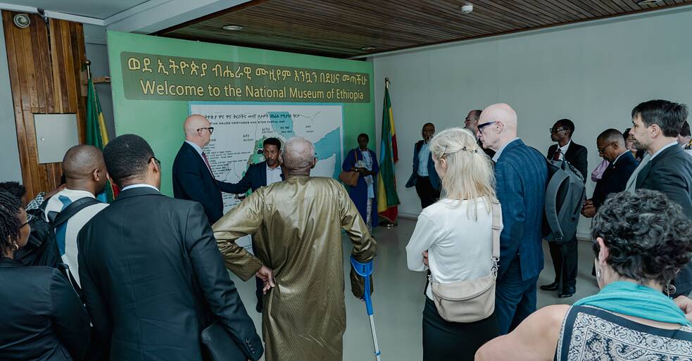 Eine Gruppe von Besuchenden versammelt sich in der Eingangshalle des Nationalmuseums von Äthiopien vor einem großen grünen Willkommensbanner in Amharisch und Englisch: „Welcome to the National Museum of Ethiopia." Ein Museumsvertreter spricht zur Gruppe neben einer Teilkarte äthiopischer Kulturerbestätten.