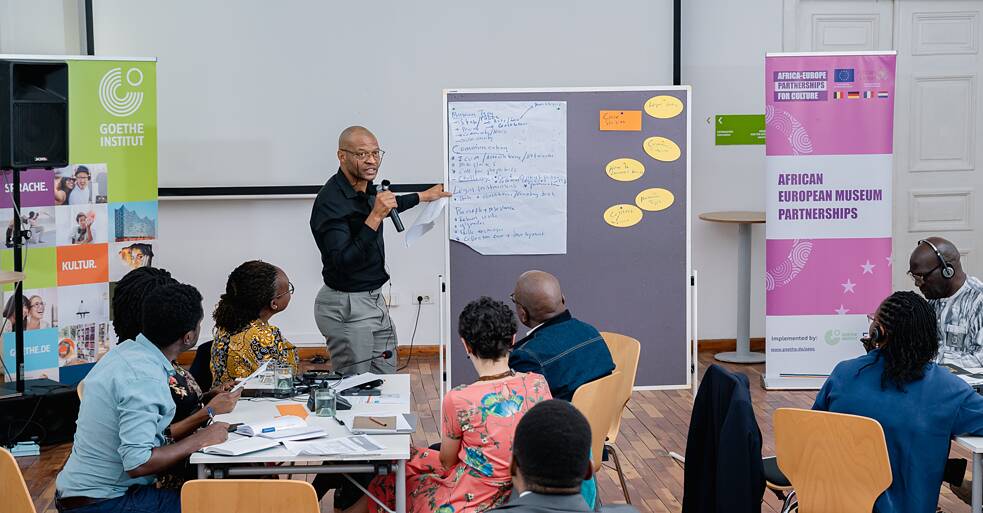 Ein Moderator steht an einer Pinnwand und präsentiert Ergebnisse vor sitzenden Teilnehmenden. Die Pinnwand zeigt geclusterte Notizen unter Überschriften wie „Museum Focus", „Communication" und „Partnership/Relevance". Branded Banner des Goethe-Instituts und der African European Museum Partnerships flankieren den Raum auf beiden Seiten.