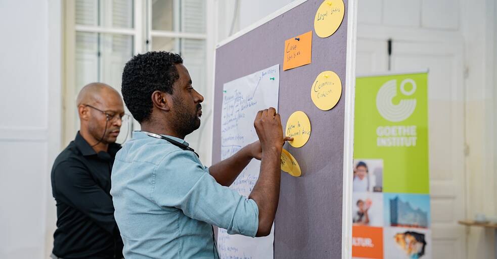 Eine Workshop-Teilnehmende heftet eine gelbe runde Karte an eine Pinnwand, während eine Kollegin von hinten zuschaut. Die Pinnwand zeigt handschriftliche Notizen auf einem Papierbogen sowie mehrere gelbe Karten mit den Beschriftungen „Legal Status", „Case Studies", „Communication" und „How to Connect/Attract". Ein Banner des Goethe-Instituts ist rechts im Bild zu sehen. Der Schauplatz ist ein heller Raum mit hoher Decke.