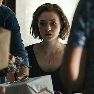 A young woman stands among a pile of presents, looking a little lost as she gazes towards the camera.