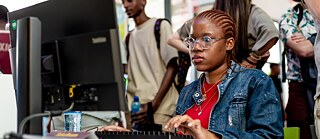 Games Connect Africa – eine Frau sitzt bei einer Veranstaltung vor einem Computer, im Hintergrund stehen Zuschauer. 