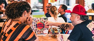 Pessoas sentadas em mesas de madeira em um ambiente de bar ou café, com destaque para duas delas observando um cardápio colorido com o título “Gira Samba” sobre a mesa. Um objeto decorativo dourado em forma de telefone também está sobre a mesa.