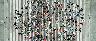 Draufsicht auf einen breiten Zebrastreifen, auf dem eine große Menge von Fußgängerinnen und Fußgängern gleichzeitig in verschiedene Richtungen die Straße überqueren.