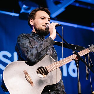 Ein Mann steht auf einer Bühne hinter einem Mikrofon. Er trägt eine Gitarre.
