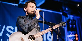 Ein Mann steht auf einer Bühne hinter einem Mikrofon. Er trägt eine Gitarre.