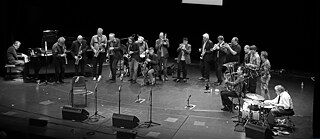 A large jazz ensemble performs on stage with brass, woodwinds, piano, and drums.