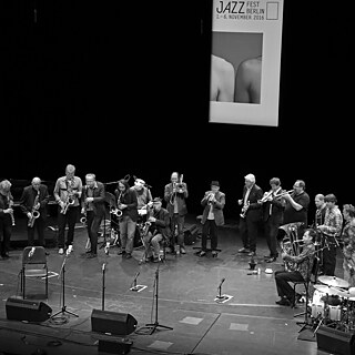 A large jazz ensemble performs on stage with brass, woodwinds, piano, and drums.