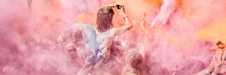 Menschenmenge bei einem Festival, umgeben von farbigem Pulver in Rosa‑, Orange‑ und Gelbtönen; eine Person wird über der Menge hochgehoben und hält die Arme nach oben.