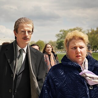 A woman and a man are looking toward the camera with serious expressions on their faces.