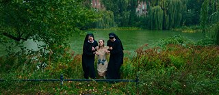 Eine junge Frau, die 2 Gewichte um ihren Bauch gebunden hat, wird von zwei Nonnen aus einem Teich gezogen