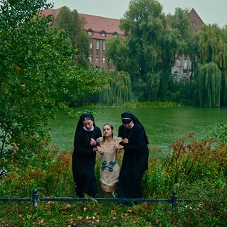Eine junge Frau, die 2 Gewichte um ihren Bauch gebunden hat, wird von zwei Nonnen aus einem Teich gezogen