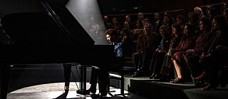 In einer dunklen Konzerthalle fällt Licht auf das Gesicht eines Pianisten, der an einem Flügel sitzt.