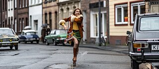 Eine junge weiße Frau springt auf einer Straße freudig in die Luft und landet mit einem Bein in einer Pfütze