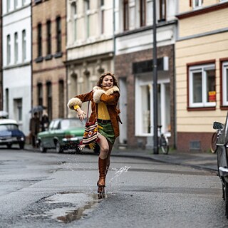 Eine junge weiße Frau springt auf einer Straße freudig in die Luft und landet mit einem Bein in einer Pfütze