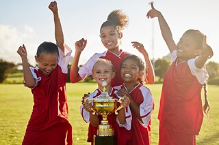 4 Mädchen in rot-weißen Fußballtrikots jubeln. Die 2 Mädchen in der Mitte tragen ein 5. Mädchen auf ihren Schultern, das einen Pokal in den Händen hält.