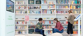 Drei Personen in einer Bibliothek mit Bücherregalen im Hintergrund, zwei sitzen auf Sitzkissen und lesen, eine steht und liest ein Buch.
