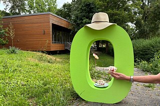 Ein grünes „O“ steht in einem Garten auf einer Wiese. Auf dem Buchstaben liegt ein Sommerhut. Eine Hand hält eine große Portion Spaghetti-Eis.