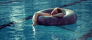Scene from the film “22 Lengths”. A person resting on a black inner tube in a blue swimming pool, one arm trailing in the water; sunlight reflects on the surface.