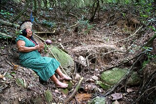 La líder indígena Iracema Kaingang se encuentra sentada junto a un manantial (Ojo del Agua) cerca del Morro do Santana, en las cercanías de Porto Alegre. A través de diversas iniciativas pedagógicas, políticas y artísticas, aboga por los derechos de los grupos indígenas y por la recuperación de la historia y la cultura de los Kaingang.
