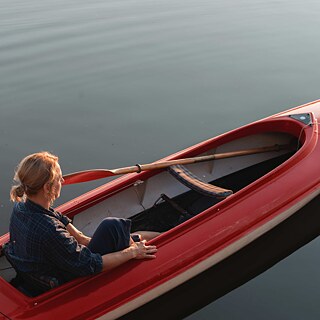 Scene from the film “Alaska”. A person sitting in a red canoe on calm water, with a paddle resting across the boat; warm light reflects on the surface.