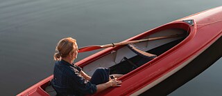 Scene from the film “Alaska”. A person sitting in a red canoe on calm water, with a paddle resting across the boat; warm light reflects on the surface.