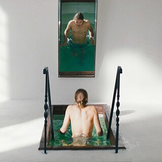 Scene from the film “Skin Deep”. A person entering a narrow indoor pool via small stairs; a vertical wall mirror reflects the upper body in the water.